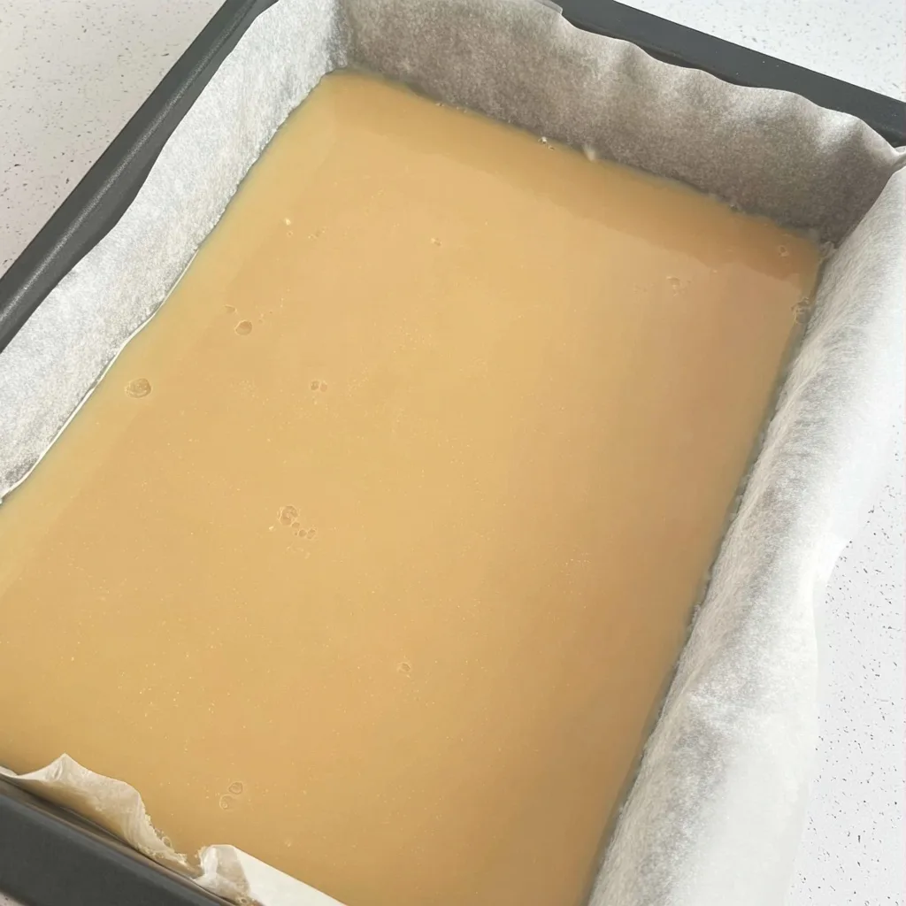 Smooth caramel layer poured over biscuit base in a baking tray lined with parchment paper