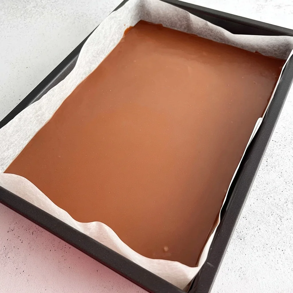 Smooth chocolate layer set on top of caramel slice in a baking tray lined with parchment paper