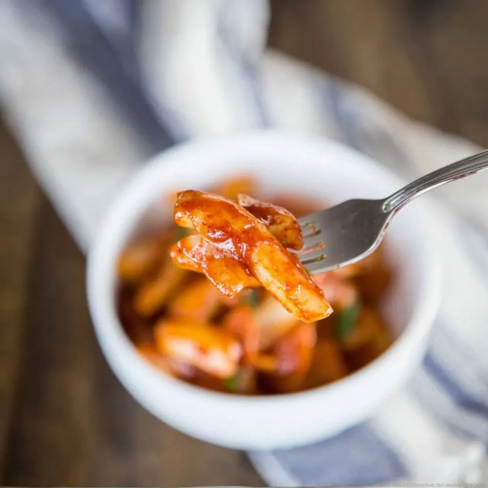 Fork holding a piece of Tteokbokki Recipe rice cake coated in spicy red sauce with a bowl of Tteokbokki Recipe in the background.
