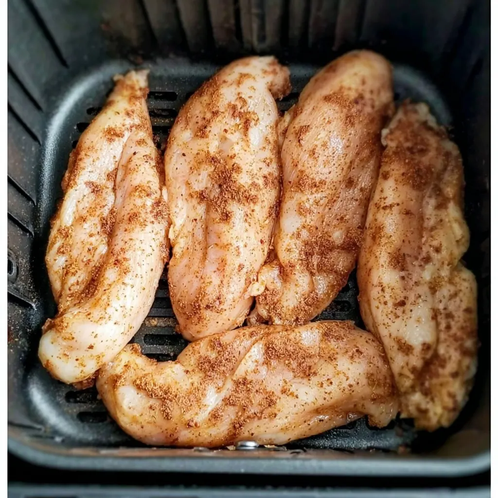 Uncooked chicken tenders seasoned with spices, arranged in an air fryer basket before starting the cooking process.