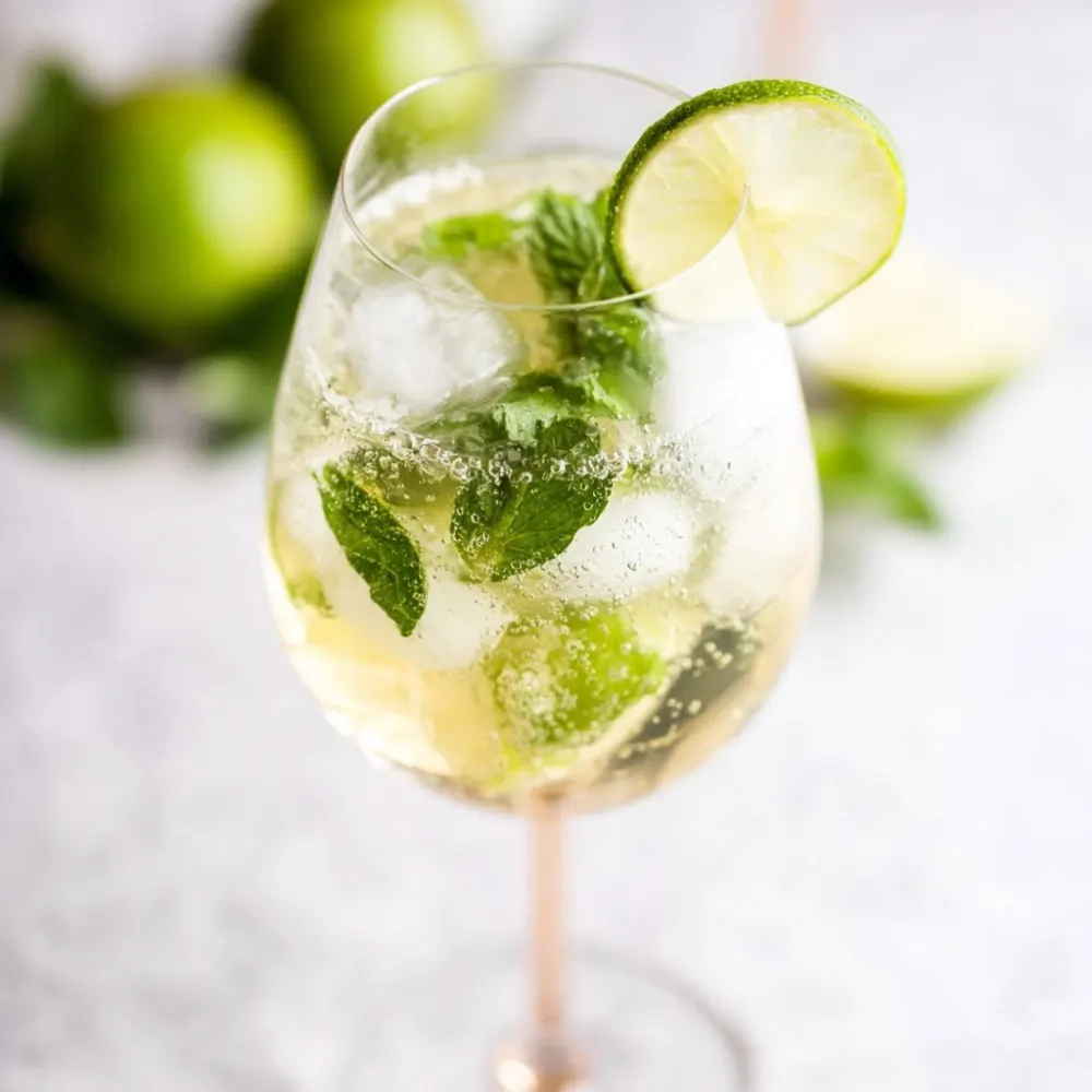 Sparkling Hugo Spritz cocktail in a tall wine glass with ice, fresh mint leaves, and lime slices, bubbles rising in the glass, with whole and halved limes blurred in the background.