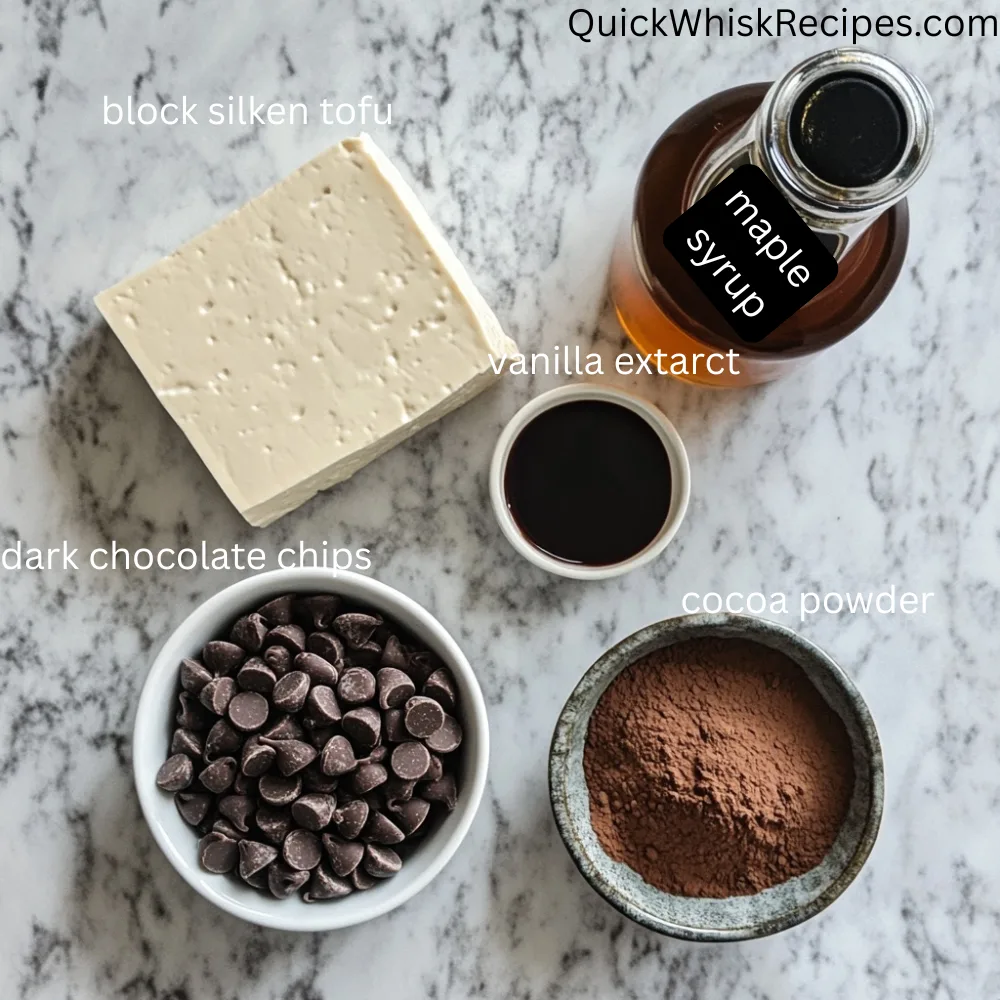 Flat lay of tofu chocolate pudding ingredients: block of silken tofu, bowl of dark chocolate chips, cocoa powder, maple syrup bottle, and vanilla extract in a small dish.