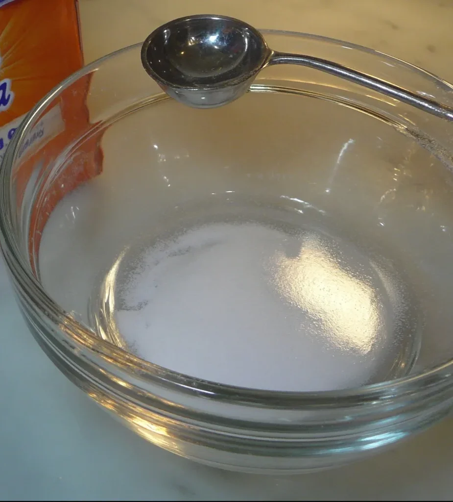 Baking soda dissolving in a glass bowl of hot water with a metal measuring spoon
