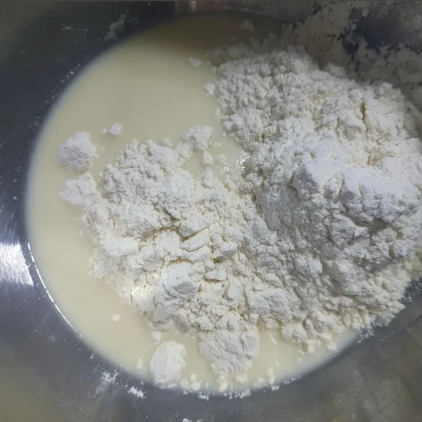 Flour being added to a bowl of milk and egg mixture, showing the beginning of pikelet batter preparation.