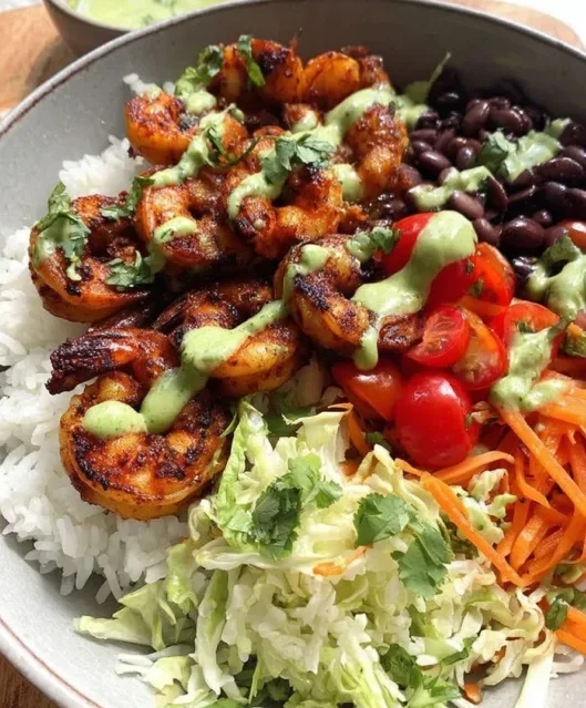 Grilled spicy shrimp bowl served with black beans, cherry tomatoes, shredded carrots, rice, and cabbage slaw, topped with creamy green sauce