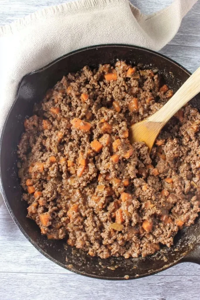 Cooked beef mince and diced carrots in a cast iron pan with a wooden spoon