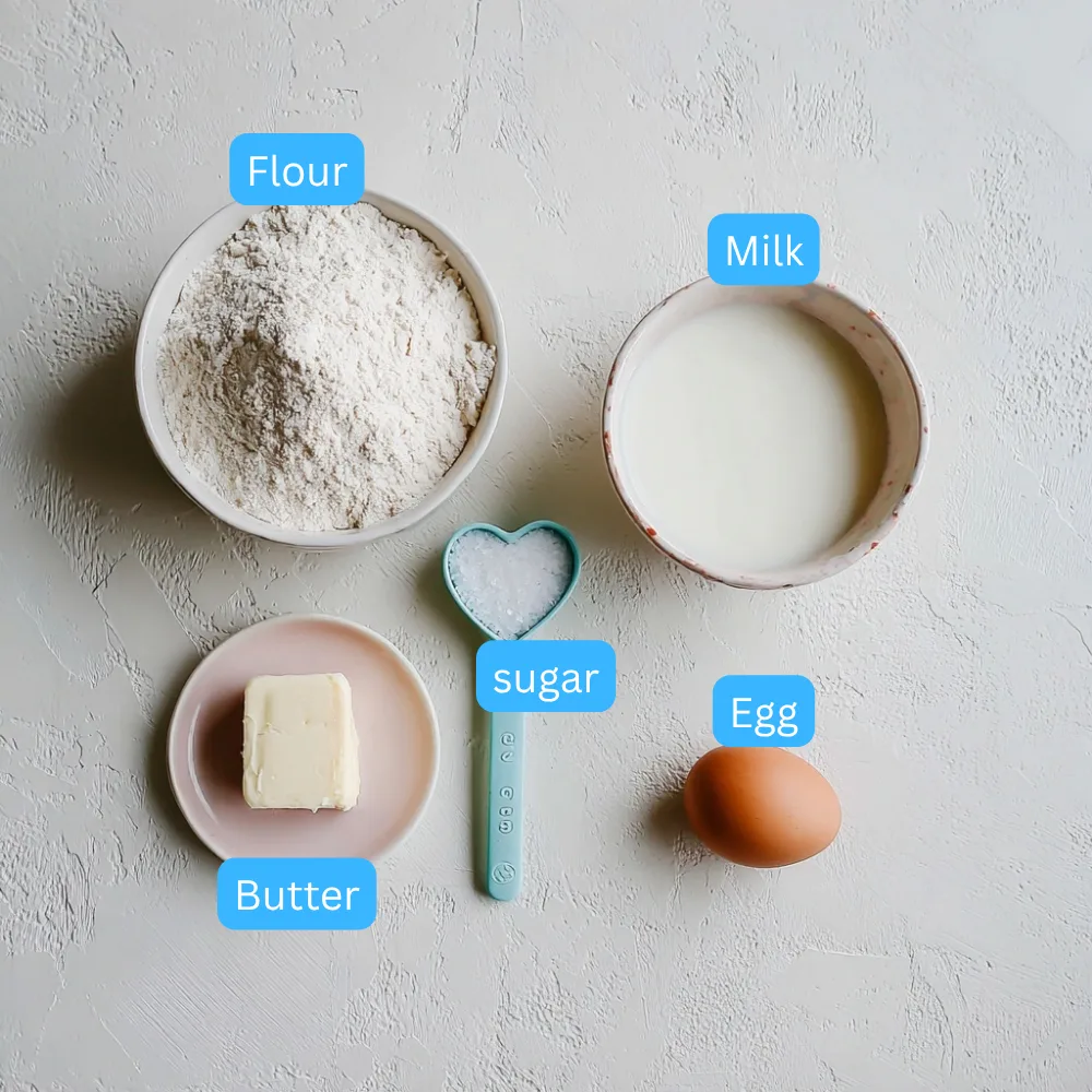 Flat lay of ingredients for pikelets including flour, milk, sugar, butter, and an egg on a light textured surface.