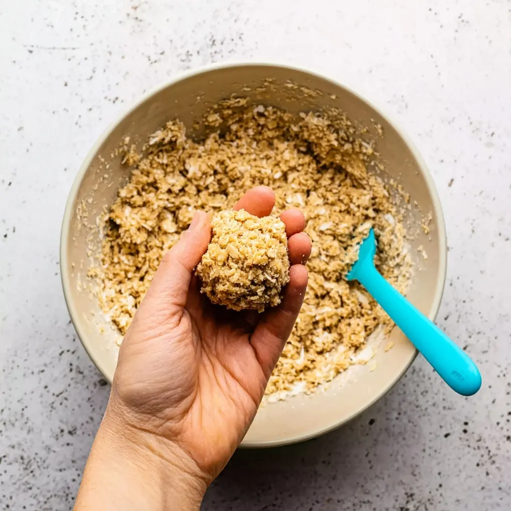 Hand holding a ball of Anzac biscuit dough above a mixing bowl with blue spatula
