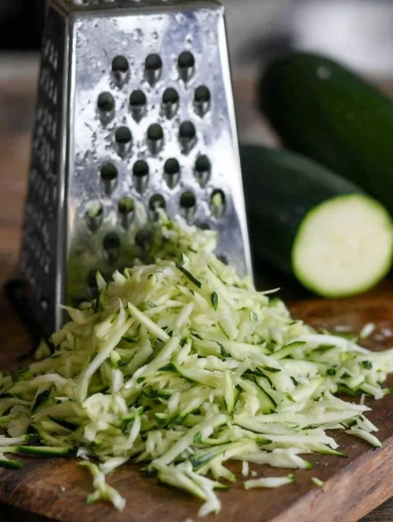 Freshly grated zucchini piled on a wooden cutting board in front of a box grater, with whole zucchinis in the background.