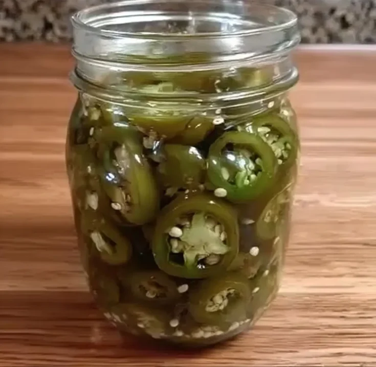 Clear mason jar filled with green pickled jalapeno slices on a wooden surface