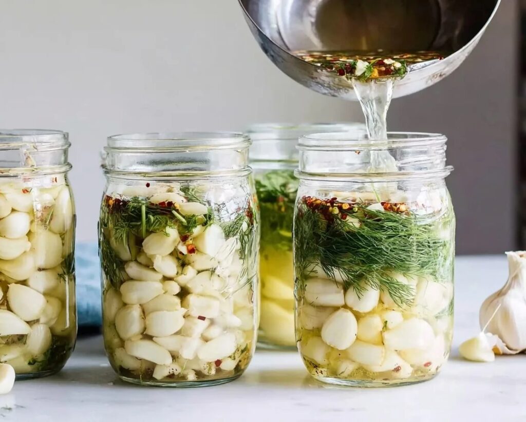 Hot vinegar brine being poured into glass jars filled with garlic cloves, dill, and chili flakes for pickling.
