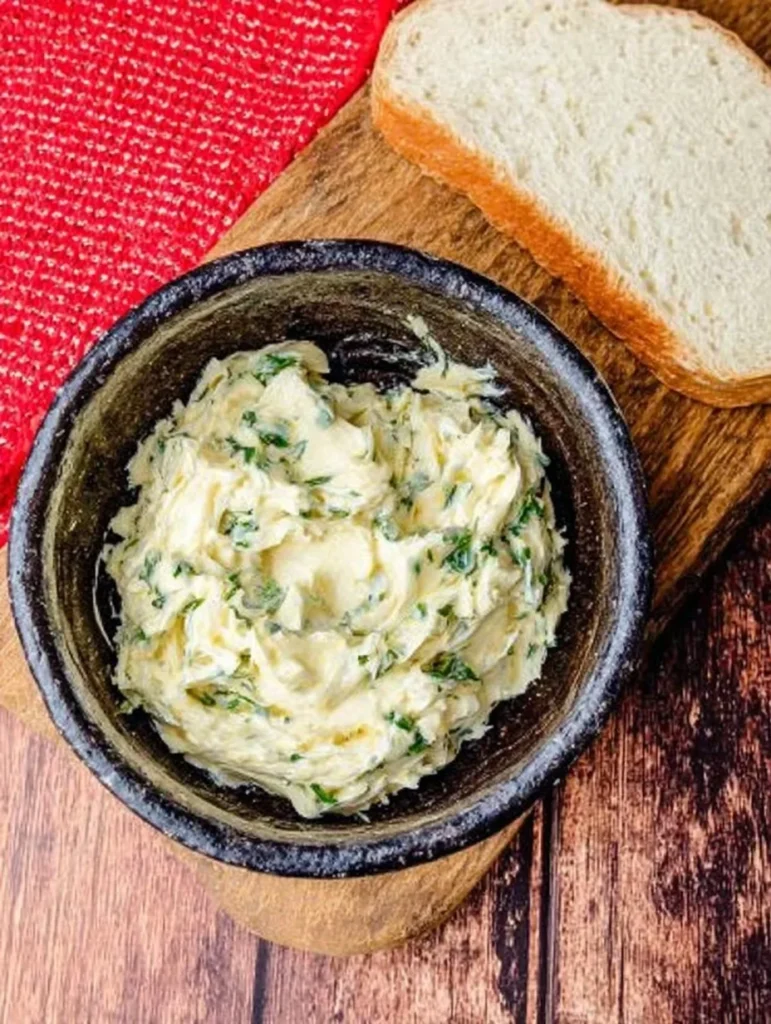 Homemade garlic butter with parsley in a bowl beside a slice of bread on wooden board