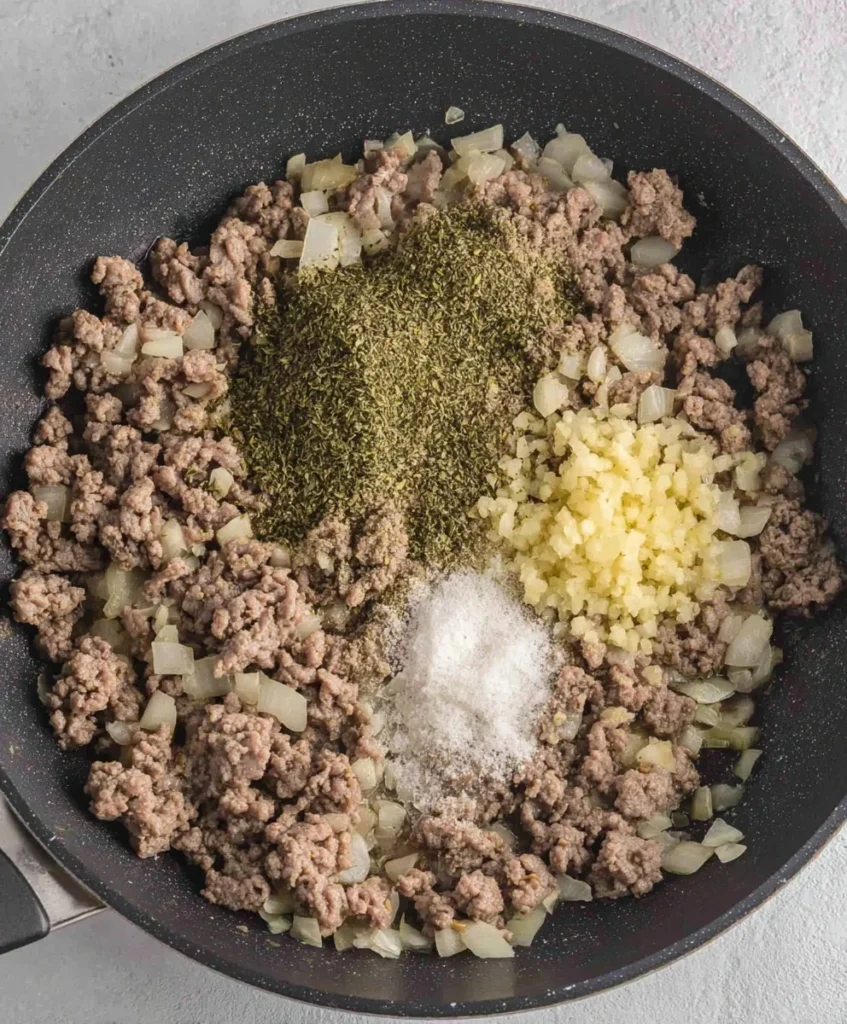 Cooked ground beef with diced onions, garlic, dried herbs, and salt in a skillet for a pasta casserole recipe.