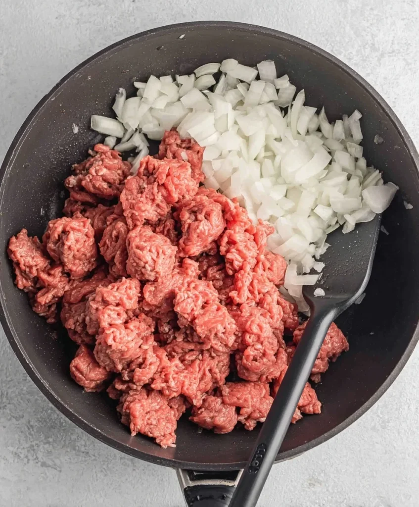 Raw ground beef with chopped onion in a skillet before browning for a hearty pasta dish.