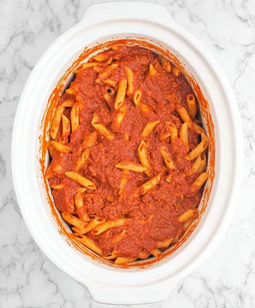 Crockpot filled with penne pasta coated in rich tomato marinara sauce during the layering process of a cheesy pasta casserole.
