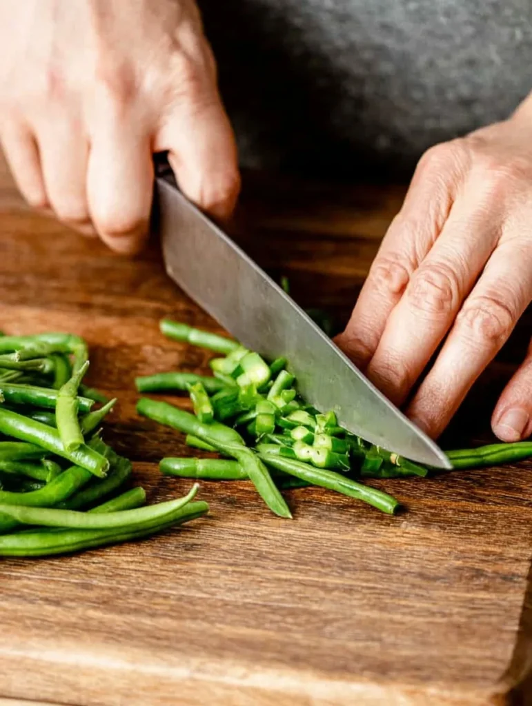 Hands using a knife to trim fresh green beans on a wooden cutting board.