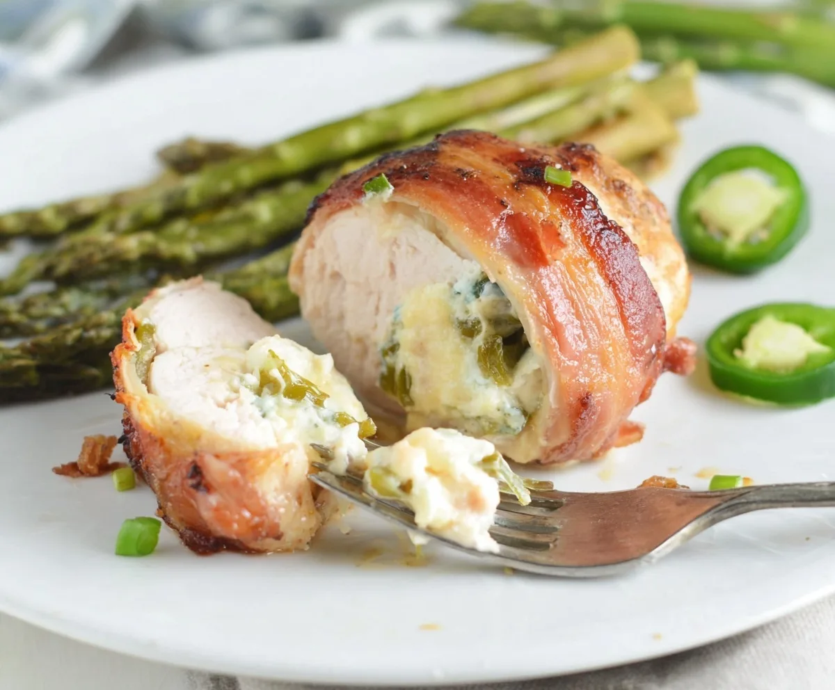 Bacon wrapped jalapeño popper chicken sliced open with creamy jalapeño cheese filling, served on a plate with roasted asparagus and fresh jalapeño slices