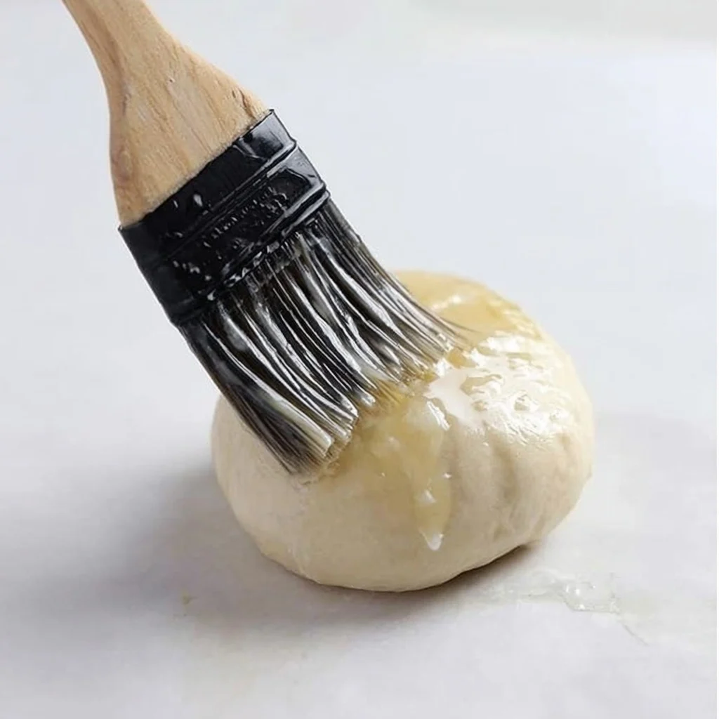 Pastry brush coating a round dough ball with melted butter before baking blueberry treats.