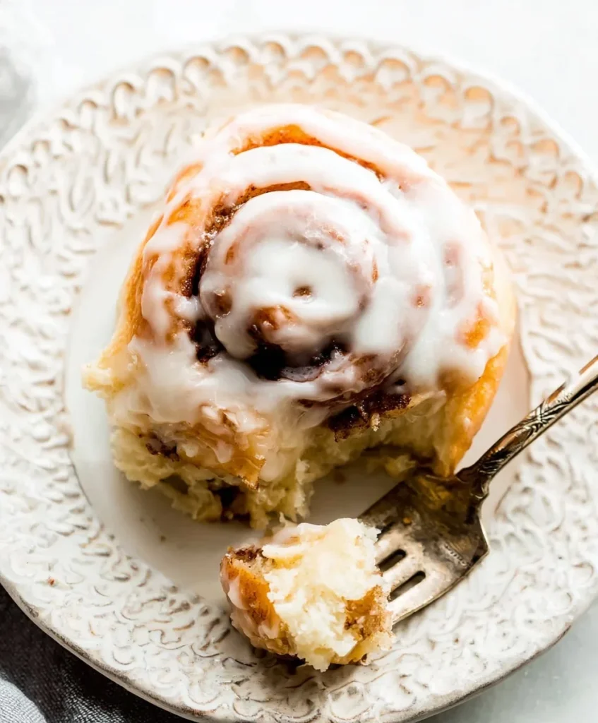 Fluffy cinnamon roll topped with cream cheese glaze served on a decorative white plate with a fork.