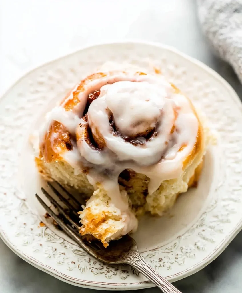 Fluffy cinnamon roll topped with vanilla glaze on a decorative white plate with a fork and a bite taken.