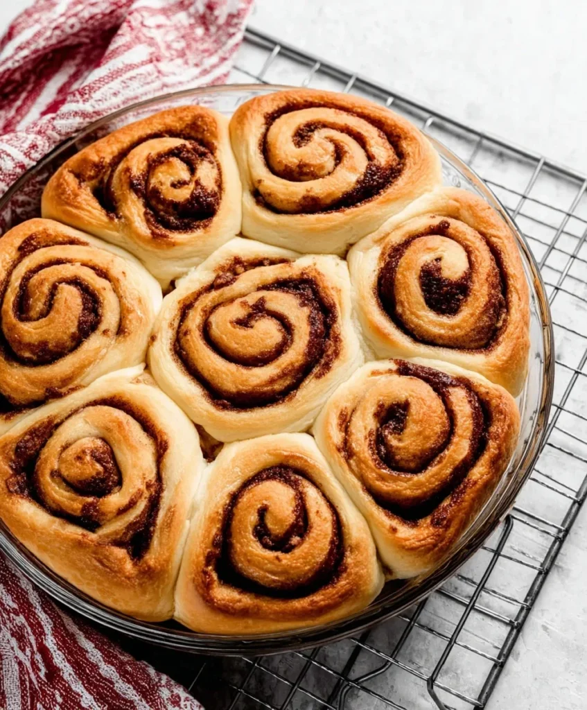 Golden brown homemade cinnamon rolls in a glass baking dish cooling on a wire rack with a red kitchen towel.