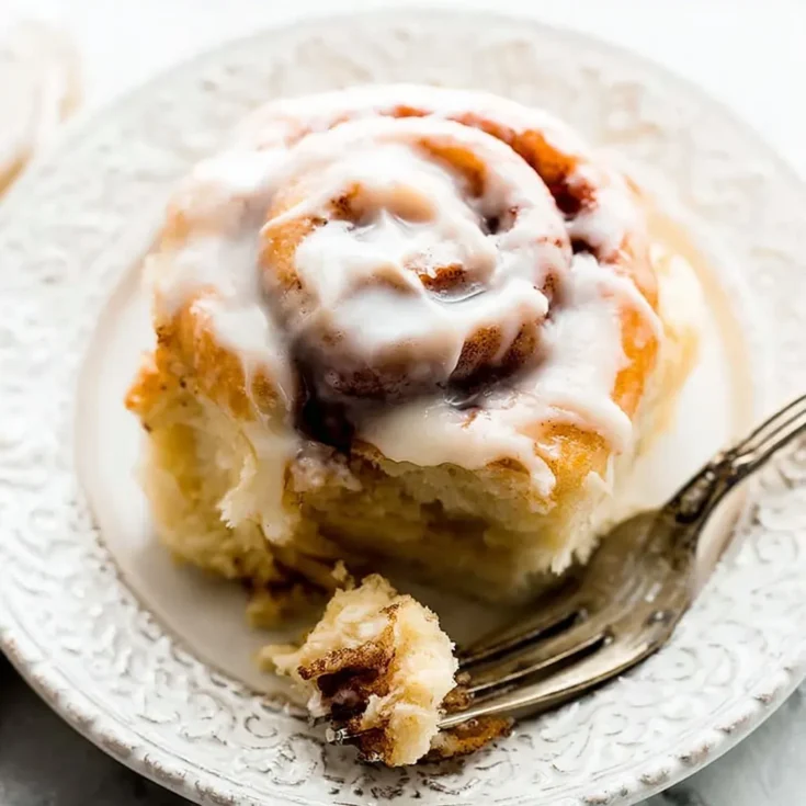 Homemade cinnamon roll covered in creamy vanilla glaze served on a white plate with a fork.