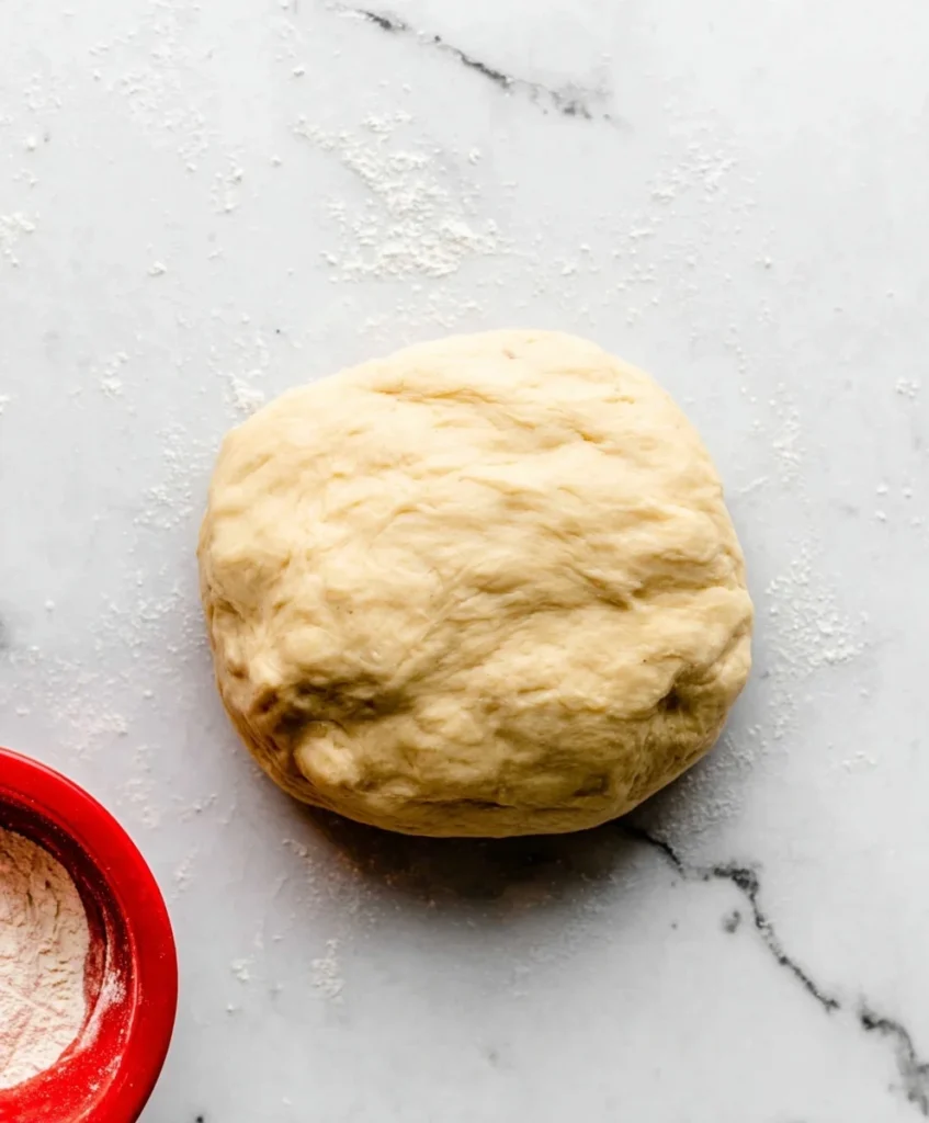 Smooth and elastic cinnamon roll dough resting on a floured marble surface with a red bowl of flour nearby.