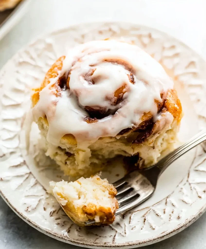 Soft cinnamon roll topped with cream cheese glaze on a white decorative plate with a fork and bite taken.