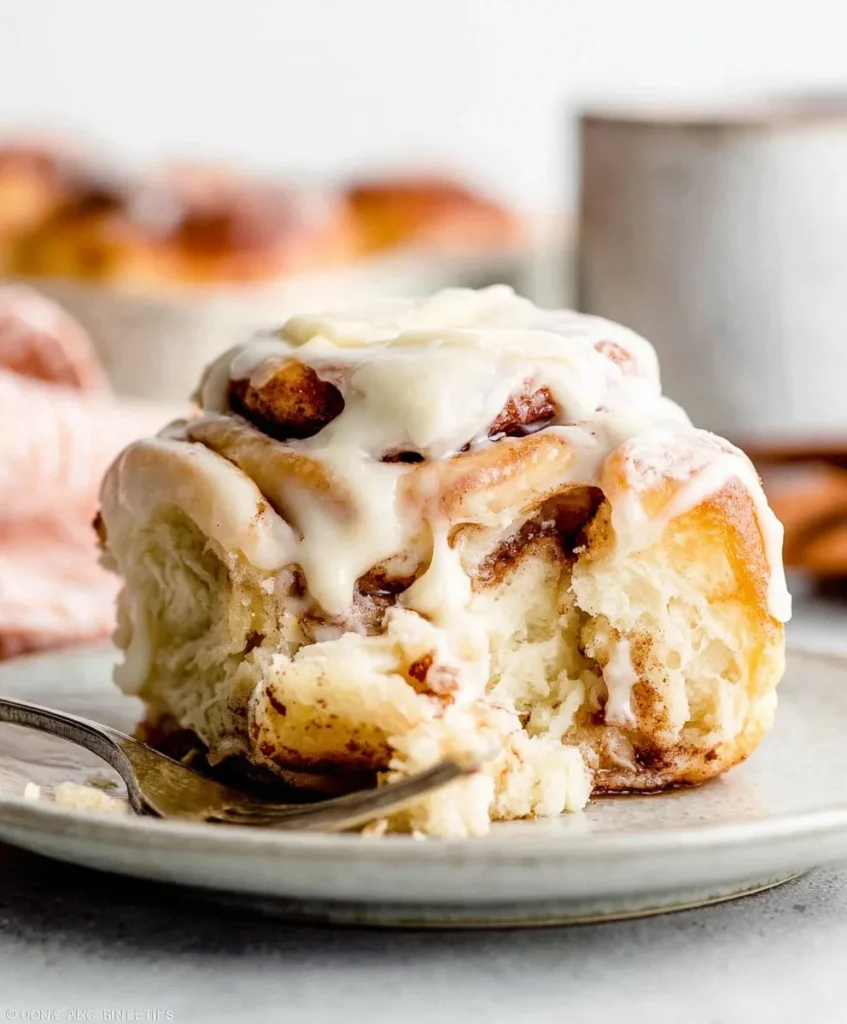 Soft homemade cinnamon roll topped with cream cheese frosting on a plate with a fork and a bite taken.