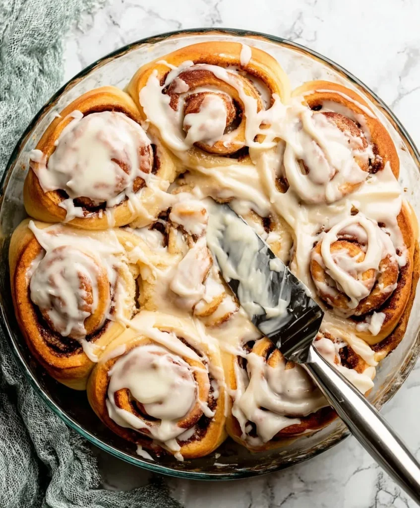 Spreading creamy frosting over freshly baked cinnamon rolls in a glass dish with a metal spatula.