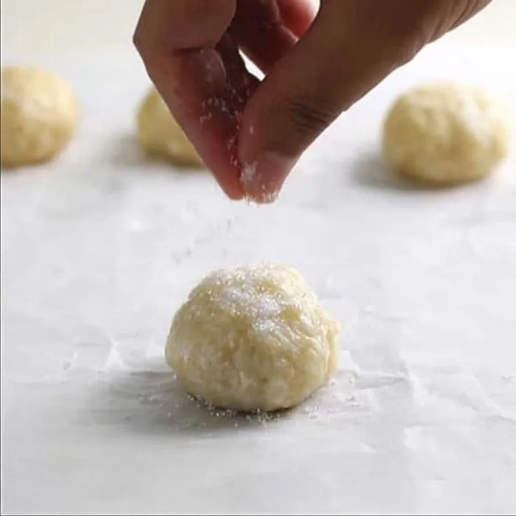 Hand sprinkling sugar over round dough balls before baking sweet blueberry pastries.