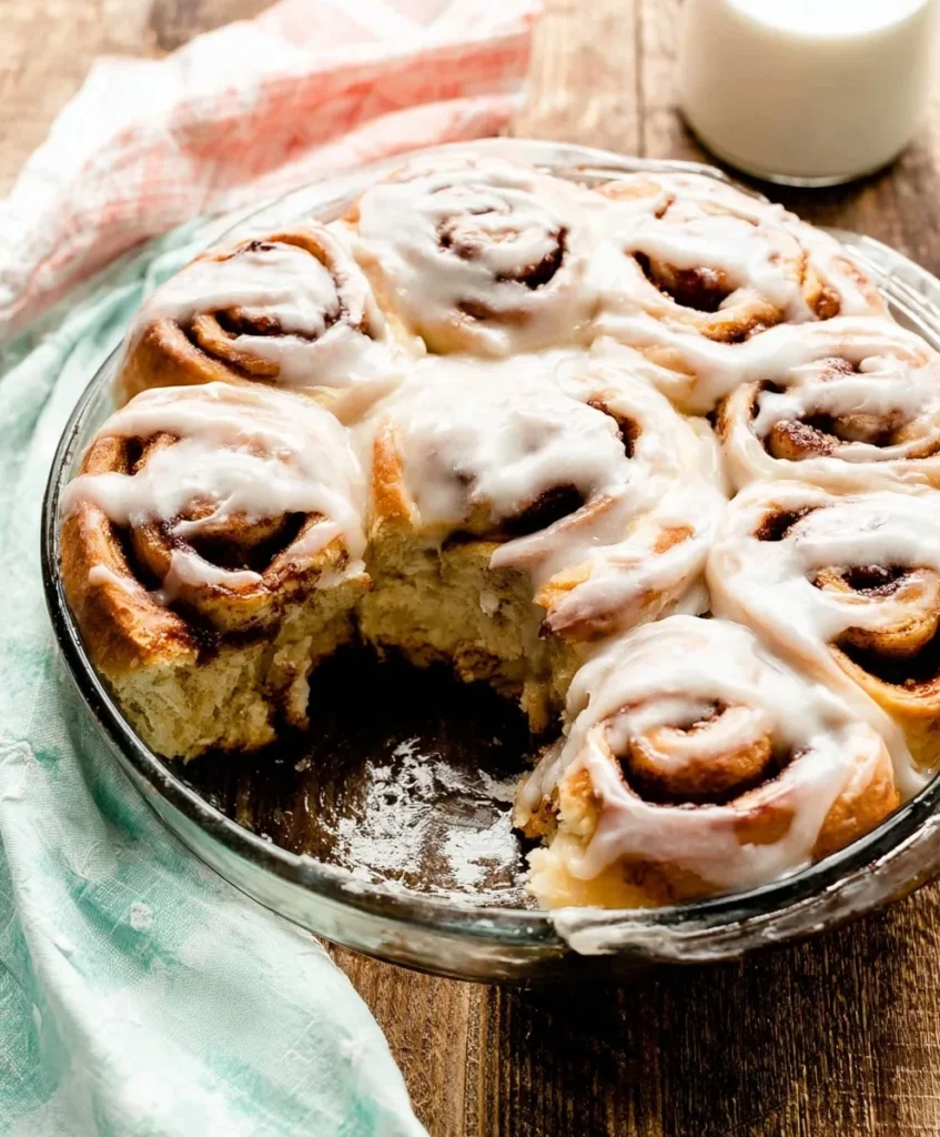Warm homemade cinnamon rolls topped with vanilla icing in a glass baking dish on a wooden table.