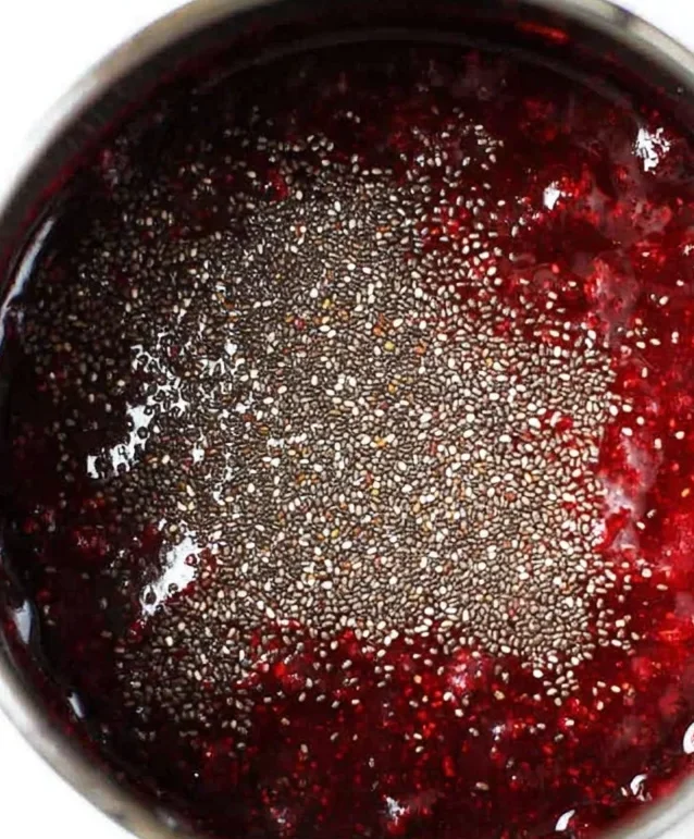 Chia seeds sprinkled over warm raspberry jam in a saucepan, showing the step where chia thickens the fruit into healthy chia seed jam.