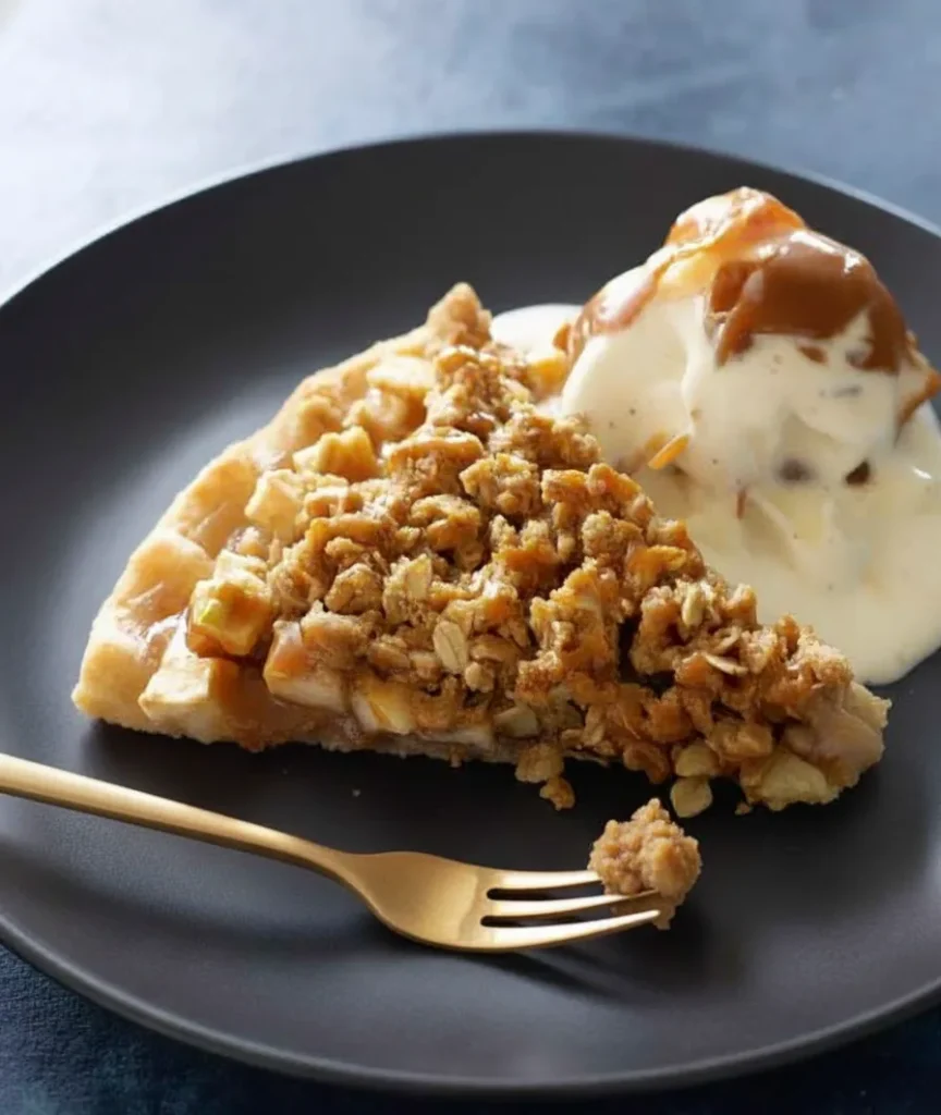 A slice of apple crisp pizza served on a black plate with a scoop of vanilla ice cream and caramel drizzle.