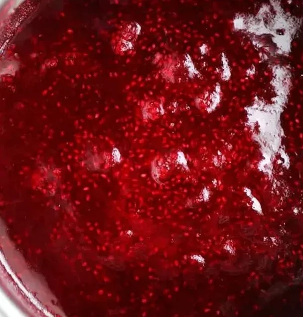 Bright red raspberry chia seed jam cooking in a saucepan, showing its thick texture and glossy surface after adding chia seeds.