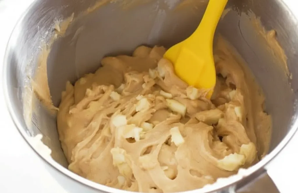 Mixing muffin batter with diced apples using a yellow spatula.