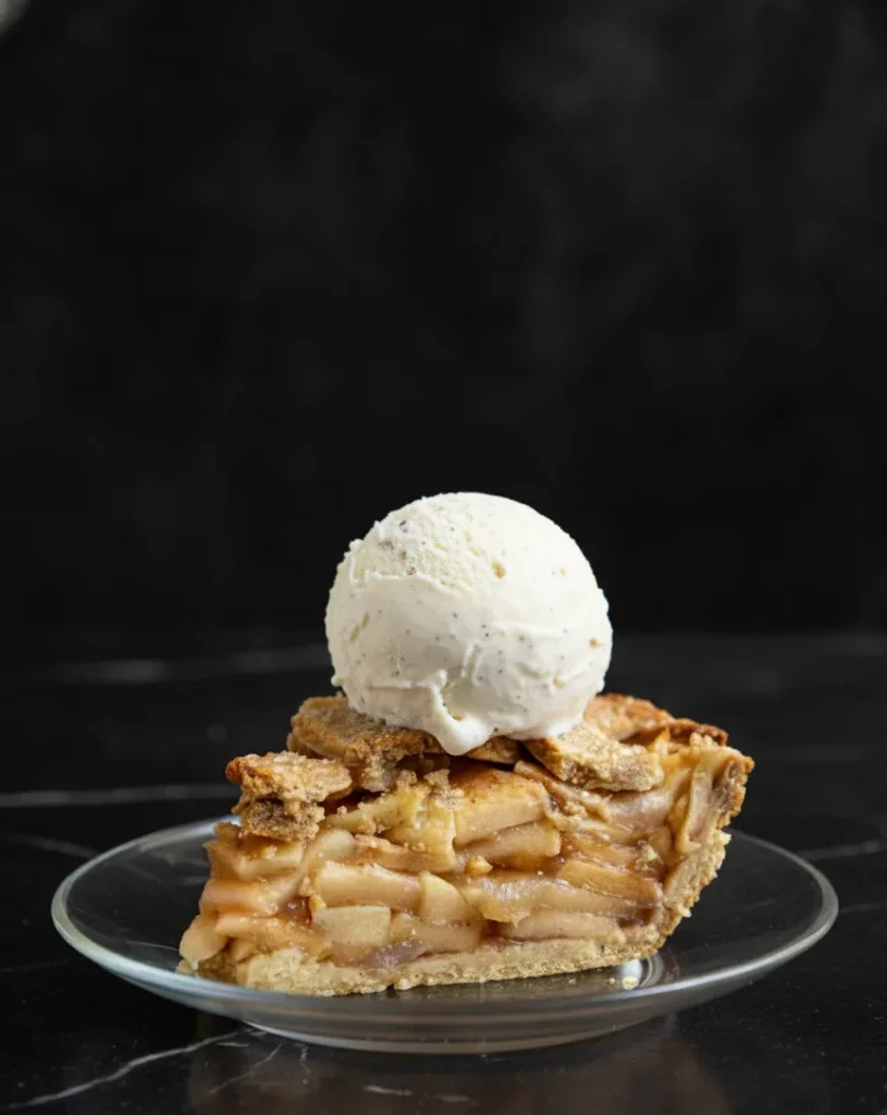 Slice of no-sugar apple pie topped with a scoop of vanilla ice cream on a glass plate.
