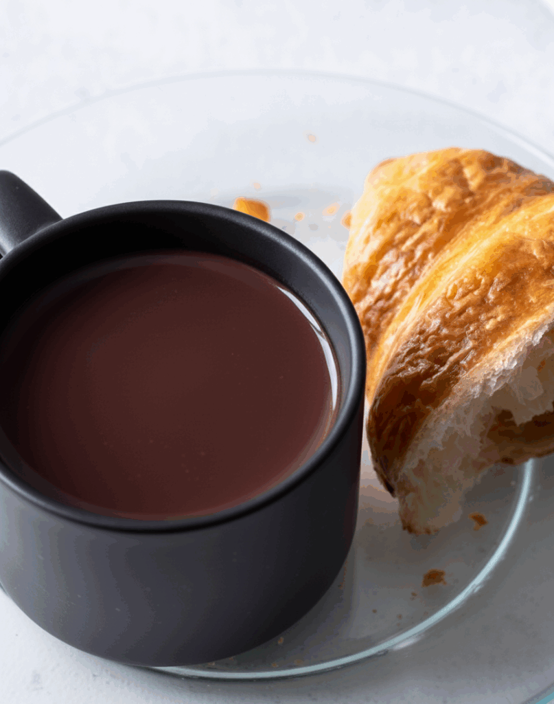 Simple French hot chocolate without cream served in a black cup with a golden croissant on a glass plate.
