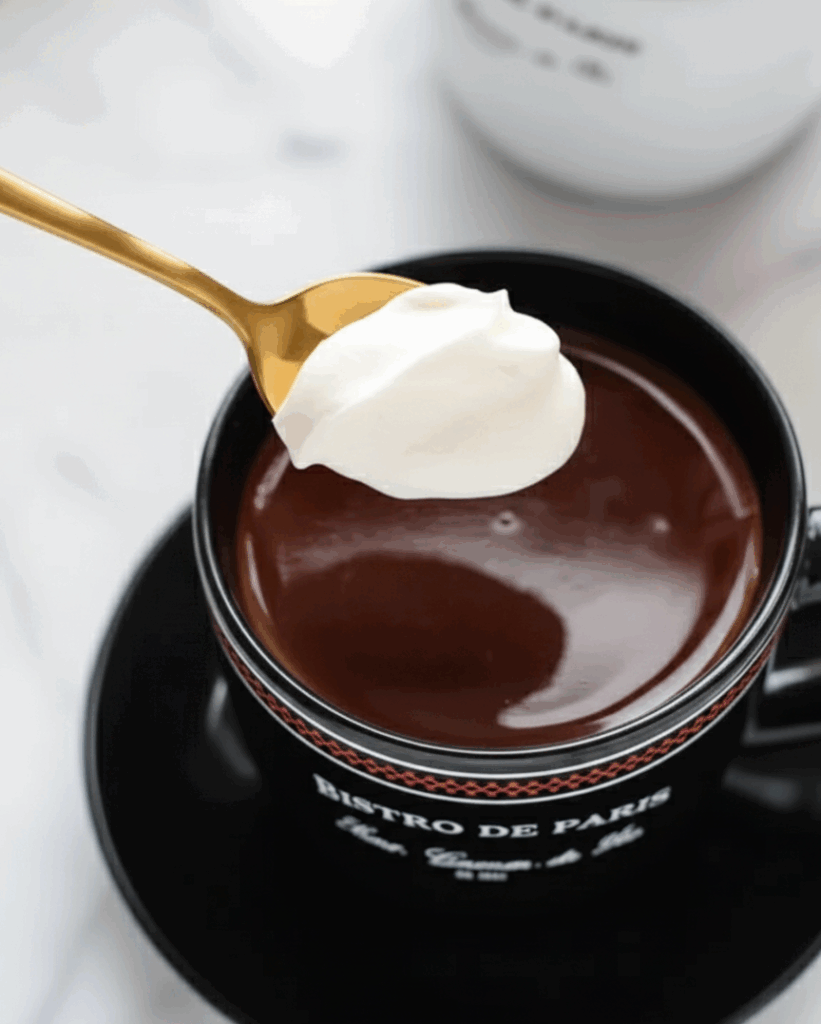 Thick Paris-style hot chocolate topped with whipped cream in a black mug with gold spoon — indulgent and creamy.