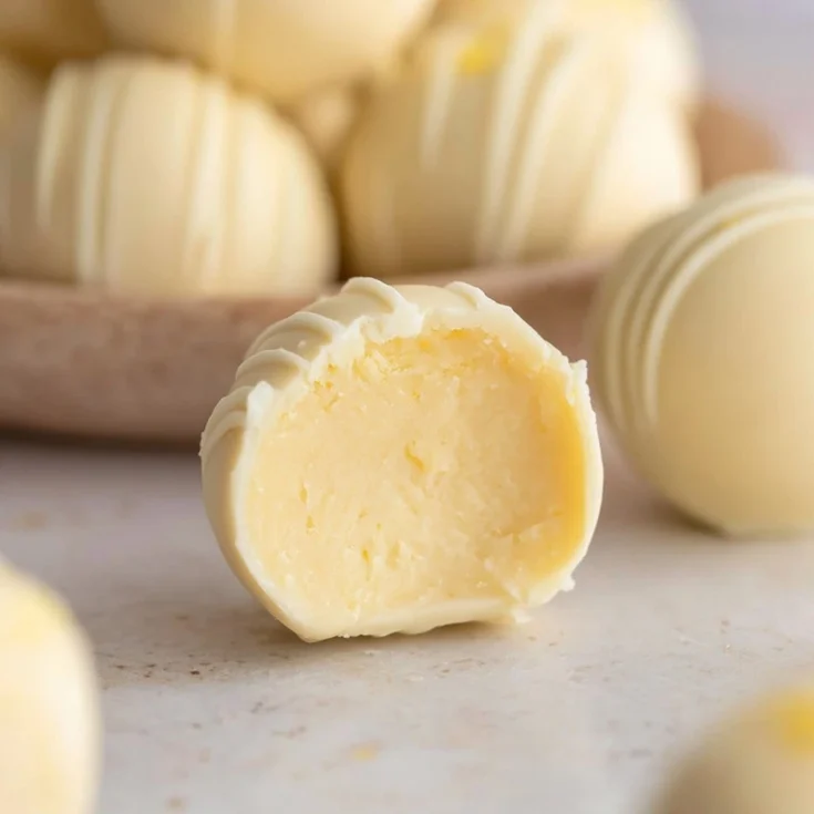 Close-up of creamy 3 ingredient lemon truffles drizzled with white chocolate and topped with fresh lemon zest on a marble serving board.