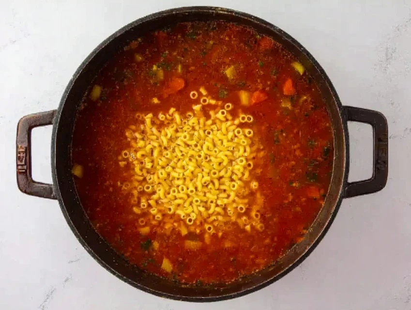 Small pasta being added to a pot of simmering minestrone soup with vegetables and tomato broth