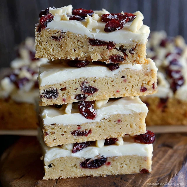 Stacked cranberry bliss bars topped with cream cheese frosting, white chocolate drizzle, and dried cranberries