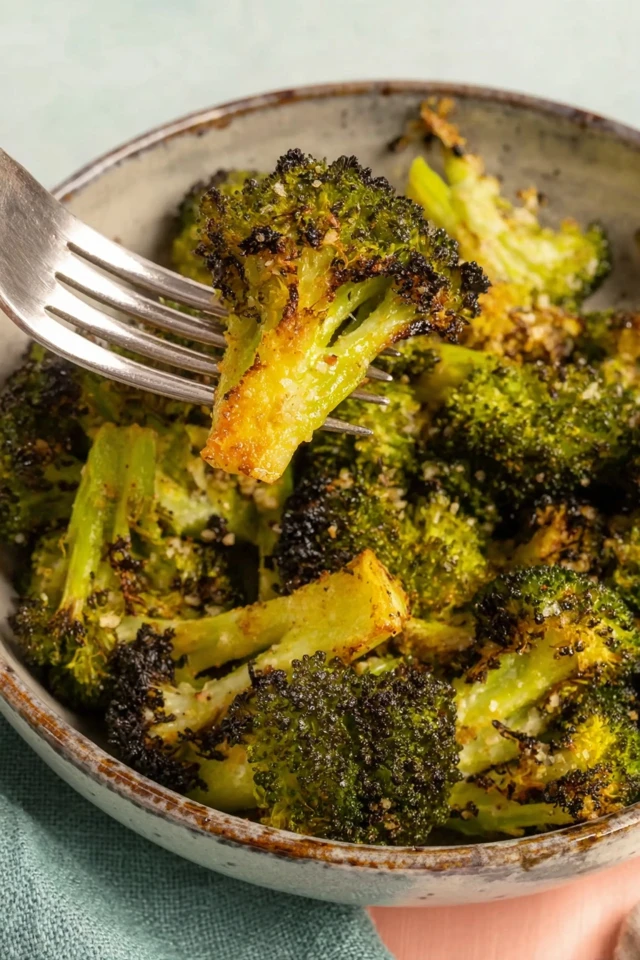 Fork lifting a piece of crispy parmesan-roasted broccoli with golden edges from a bowl