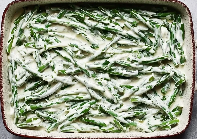Green beans mixed with creamy sauce spread evenly in a baking dish, ready for green bean casserole.