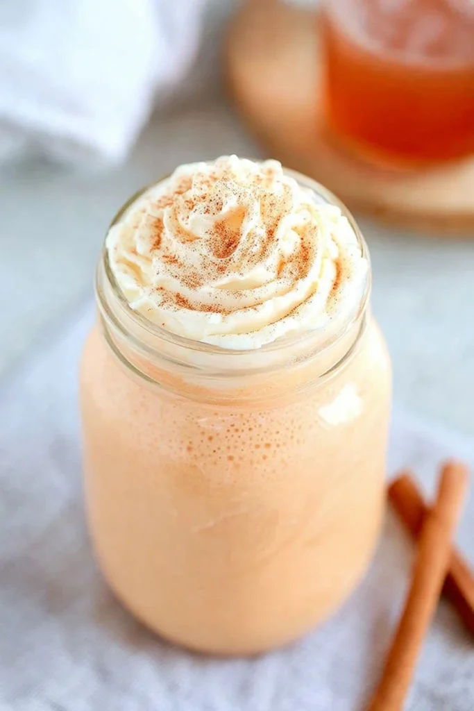 A close-up of creamy pumpkin spice milk in a mason jar topped with whipped cream and cinnamon, with cinnamon sticks and a blurred drink in the background.