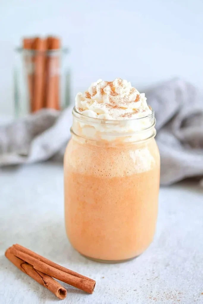 A creamy pumpkin spice milk in a mason jar topped with whipped cream and cinnamon, with cinnamon sticks and a soft cloth in the background.