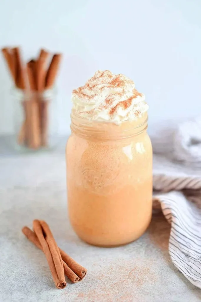 A mason jar filled with creamy pumpkin spice milk topped with whipped cream and cinnamon, with cinnamon sticks and a striped cloth on a light surface.