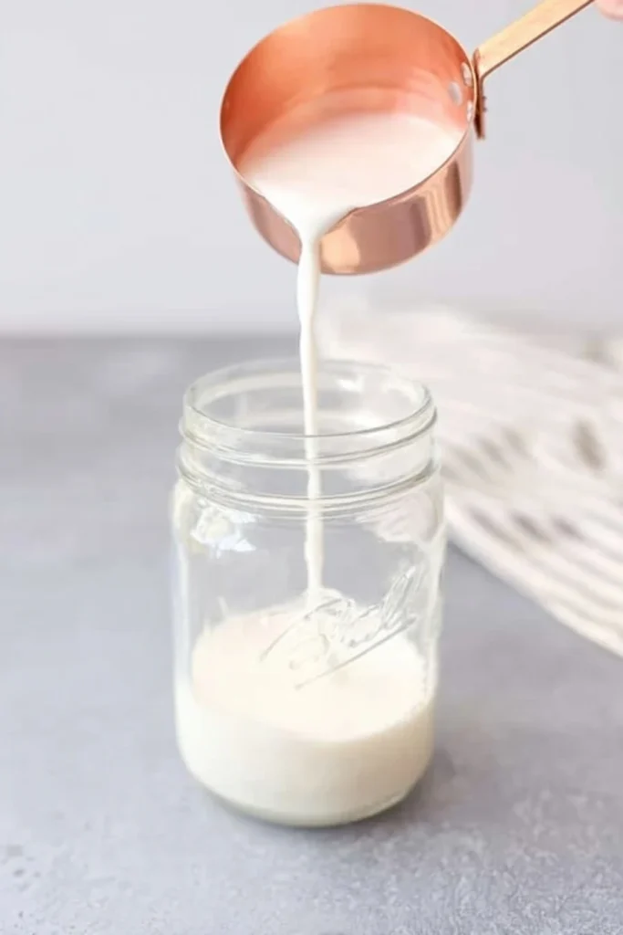 Milk being poured from a copper measuring cup into a glass jar on a gray surface.