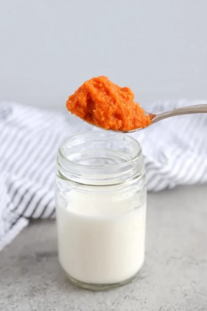 A spoonful of pumpkin purée being added to a jar of milk on a light gray surface with a striped cloth in the background.