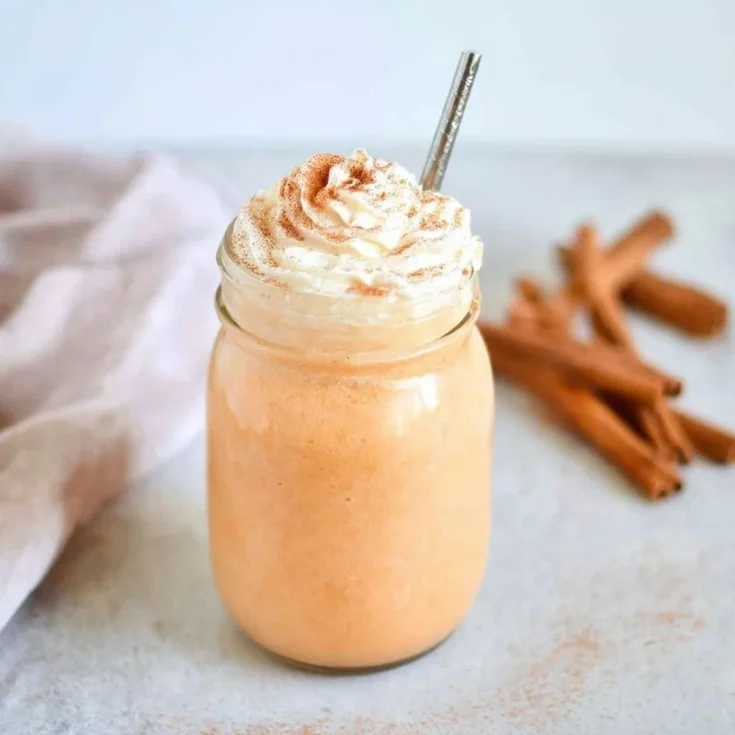 A mason jar filled with creamy pumpkin spice milk topped with whipped cream and cinnamon, with cinnamon sticks in the background.